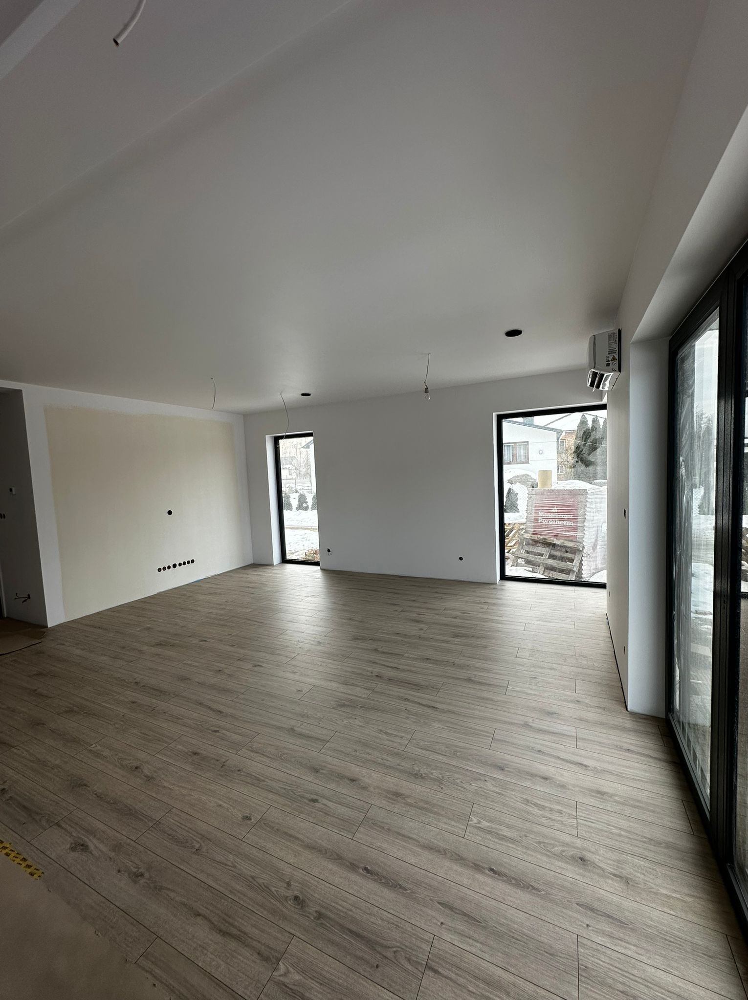 Jasne panele podłogowe w przestronnym, surowym wnętrzu w Jarosławiu. Widoczne przygotowane ściany do malowania i duże okna z czarnymi ramami. Minimalistyczny styl.