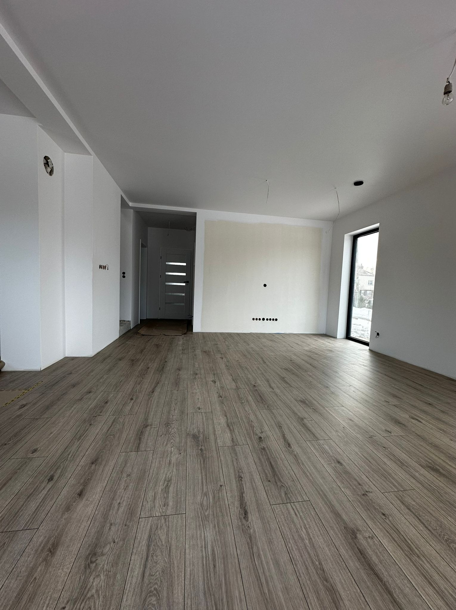 Jasne panele podłogowe w przestronnym, surowym wnętrzu z białymi ścianami i dużym oknem. Widoczne przygotowania do wykończenia, wystające kable.