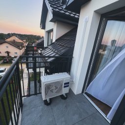 KLIMATYZACJA Michał Dudek - Klimatyzator Haier na balkonie z czarną balustradą, na tle zabudowy osiedlowej i zachodzącego słońca, widoczny fragment okna z firanką.
