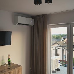 KLIMATYZACJA Michał Dudek - Nowoczesny pokój z klimatyzatorem Haier na ścianie, widok na balkon z jednostką zewnętrzną klimatyzacji, butelka szampana na komodzie, oświetlenie sufitowe.