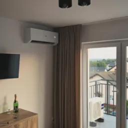 Nowoczesny pokój z klimatyzatorem Haier na ścianie, widok na balkon z jednostką zewnętrzną klimatyzacji, butelka szampana na komodzie, oświetlenie sufitowe.