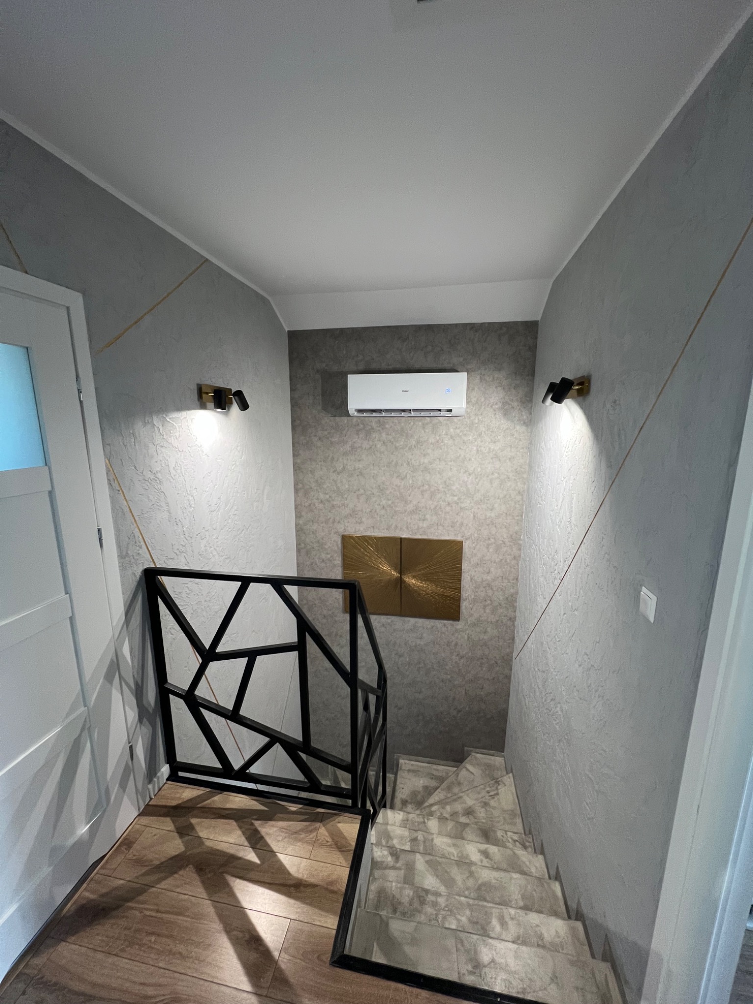 Korytarz z klimatyzatorem zamontowanym nad schodami, nowoczesna balustrada z czarnych profili, ściany z dekoracyjnym tynkiem i złotymi akcentami, oświetlenie punktowe.