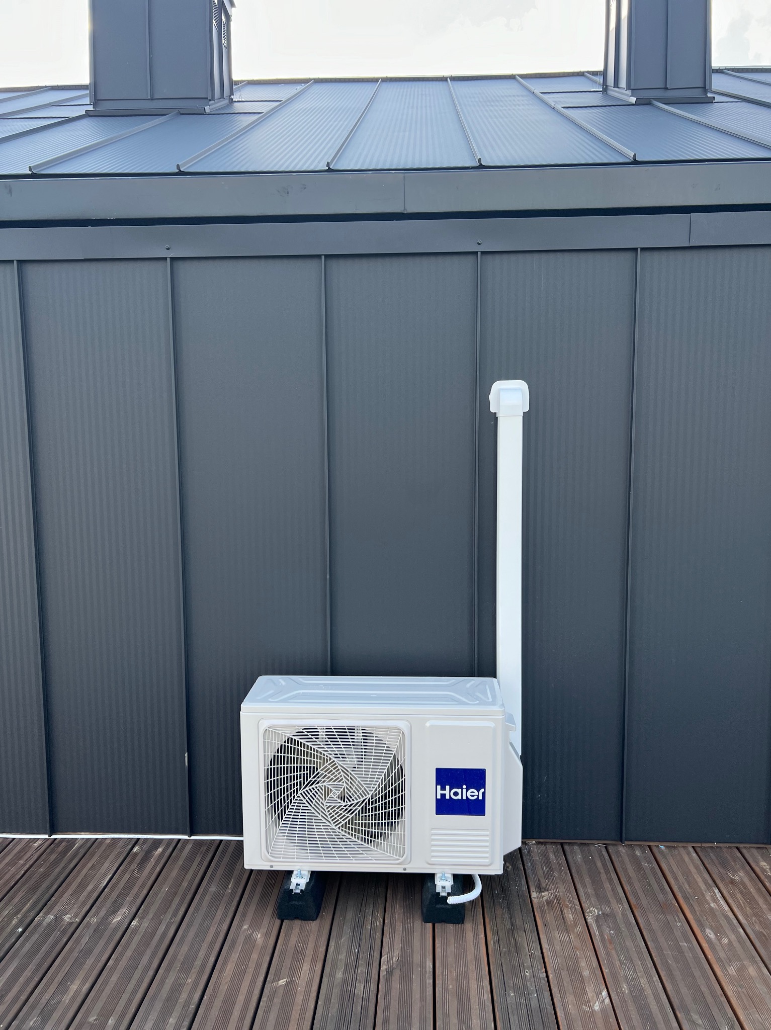 Jednostka zewnętrzna klimatyzacji Haier na drewnianym tarasie w Sławnie, zamontowana na czarnych podkładkach antywibracyjnych, z szarym, pionowo żłobkowanym tłem ściany budynku.
