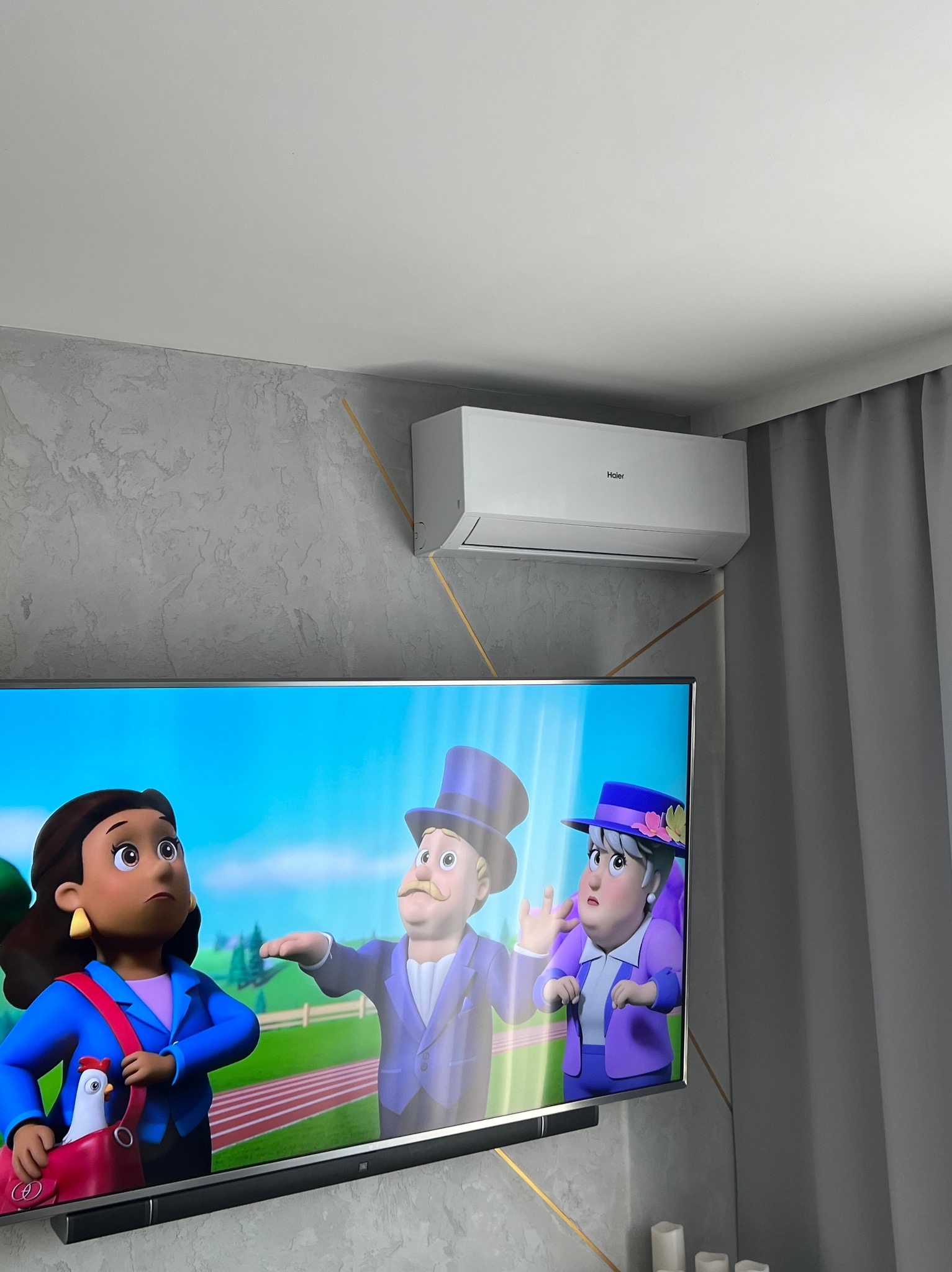 Biały klimatyzator Haier zamontowany na ścianie z dekoracyjnym wzorem, widoczny fragment telewizora z kreskówką.
