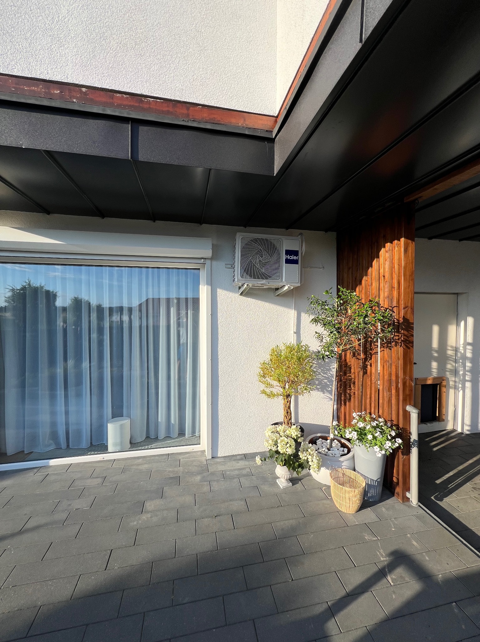 Biała jednostka zewnętrzna klimatyzacji Haier zamontowana na ścianie budynku, obok okna z roletą i drewnianej kolumny, z doniczkami z kwiatami na szarym bruku.