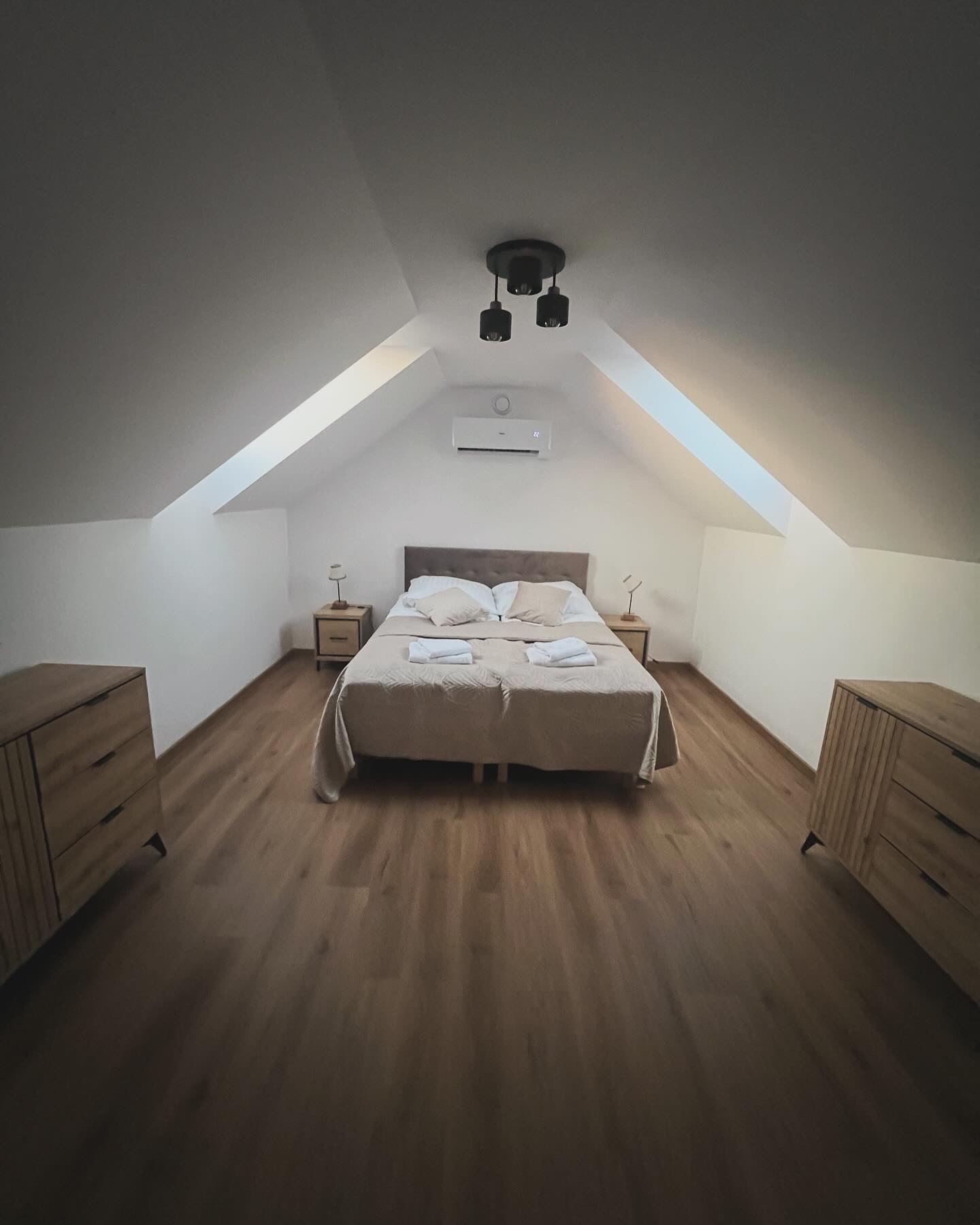 Pokój na poddaszu z drewnianą podłogą, łóżkiem małżeńskim, dwiema szafkami nocnymi i komodami po bokach, oświetlony lampą sufitową i naturalnym światłem z okna dachowego, z zamontowanym...