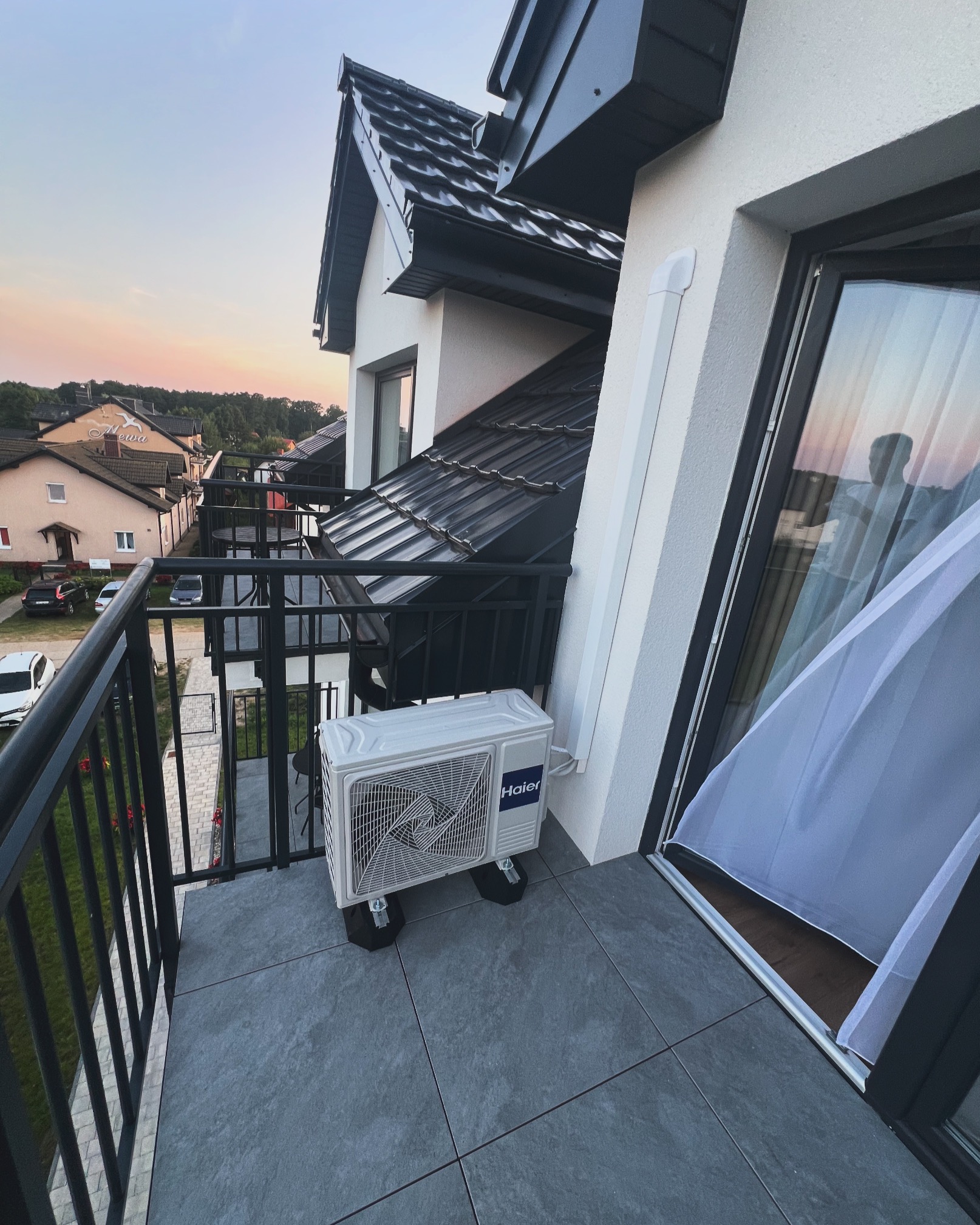 Klimatyzator Haier na balkonie z czarną balustradą, na tle zabudowy osiedlowej i zachodzącego słońca, widoczny fragment okna z firanką.