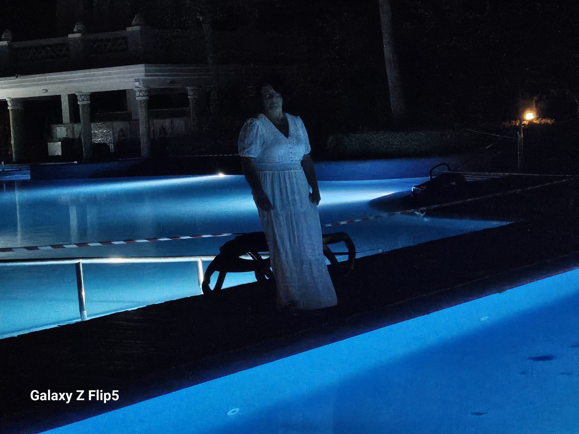 Kobieta w białej sukni stoi na pomoście przy oświetlonym basenie nocą. W tle widoczny budynek z kolumnami i lampa oświetlająca teren.