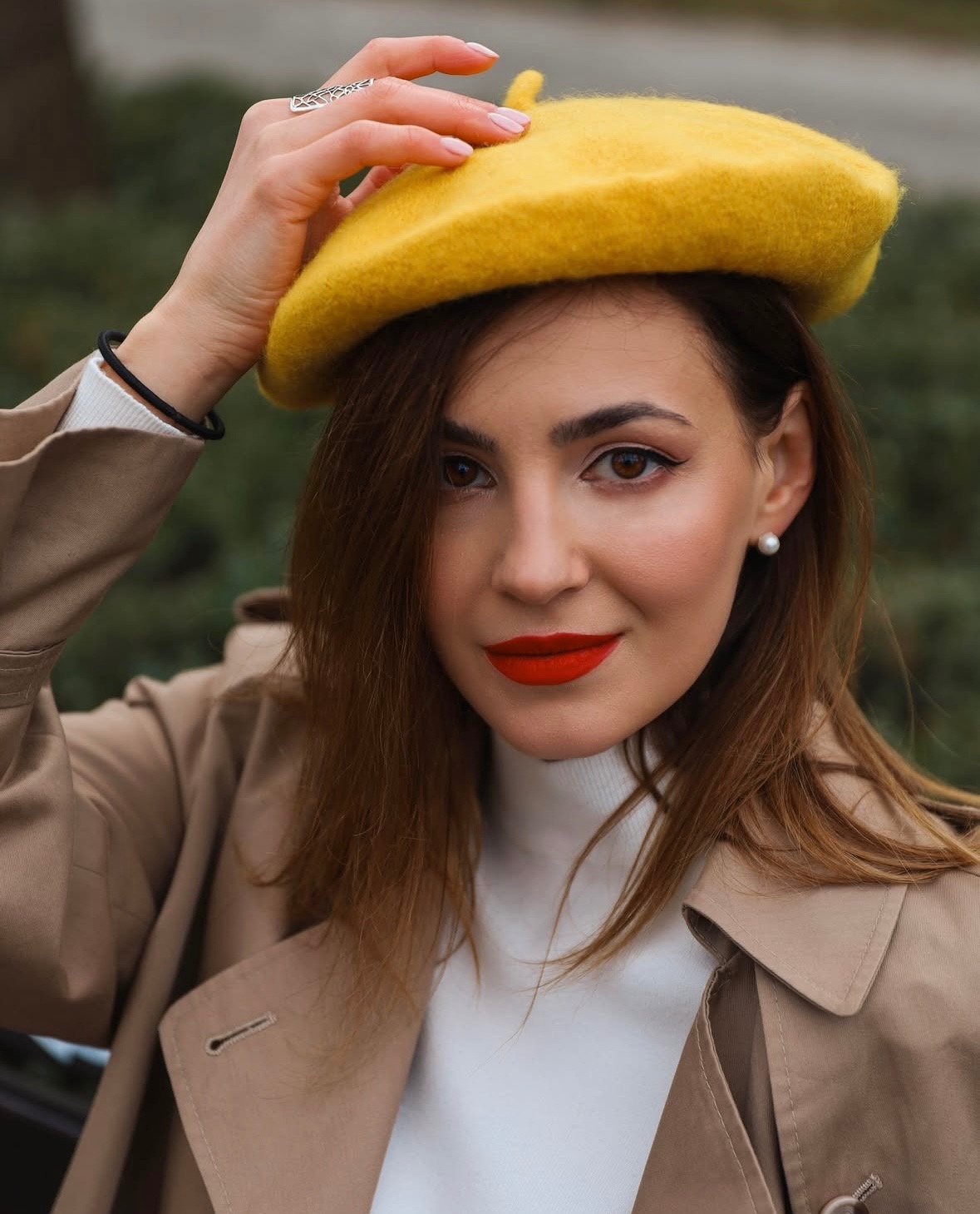 Portret kobiety w beżowym płaszczu i żółtym berecie, z czerwoną szminką i perłą w uchu, trzymającej beret dłonią z ozdobnym pierścionkiem.