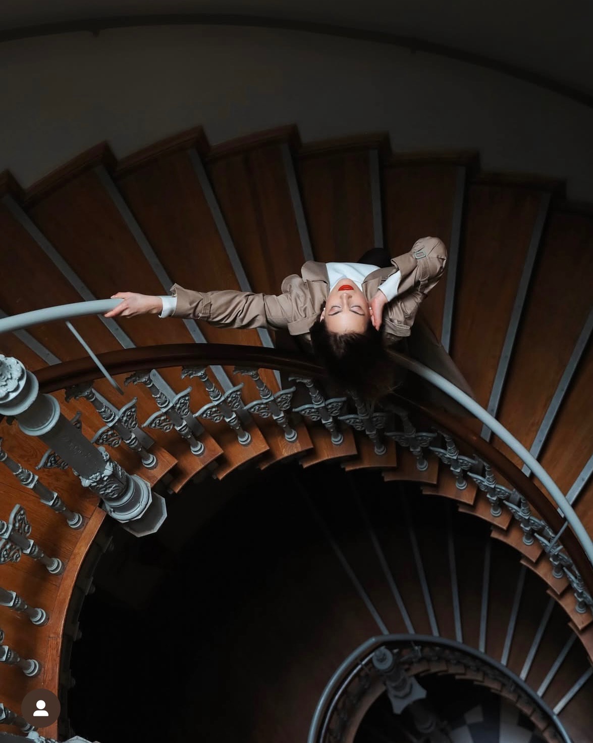 Kobieta w beżowym żakiecie leży na krętych, drewnianych schodach z metalową balustradą, ujęcie z góry.