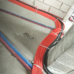 Wiązka pomarańczowych i czarnych rur instalacyjnych, przymocowanych metalowymi obejmami do surowego podłoża, biegnąca wzdłuż ściany z bloczków.