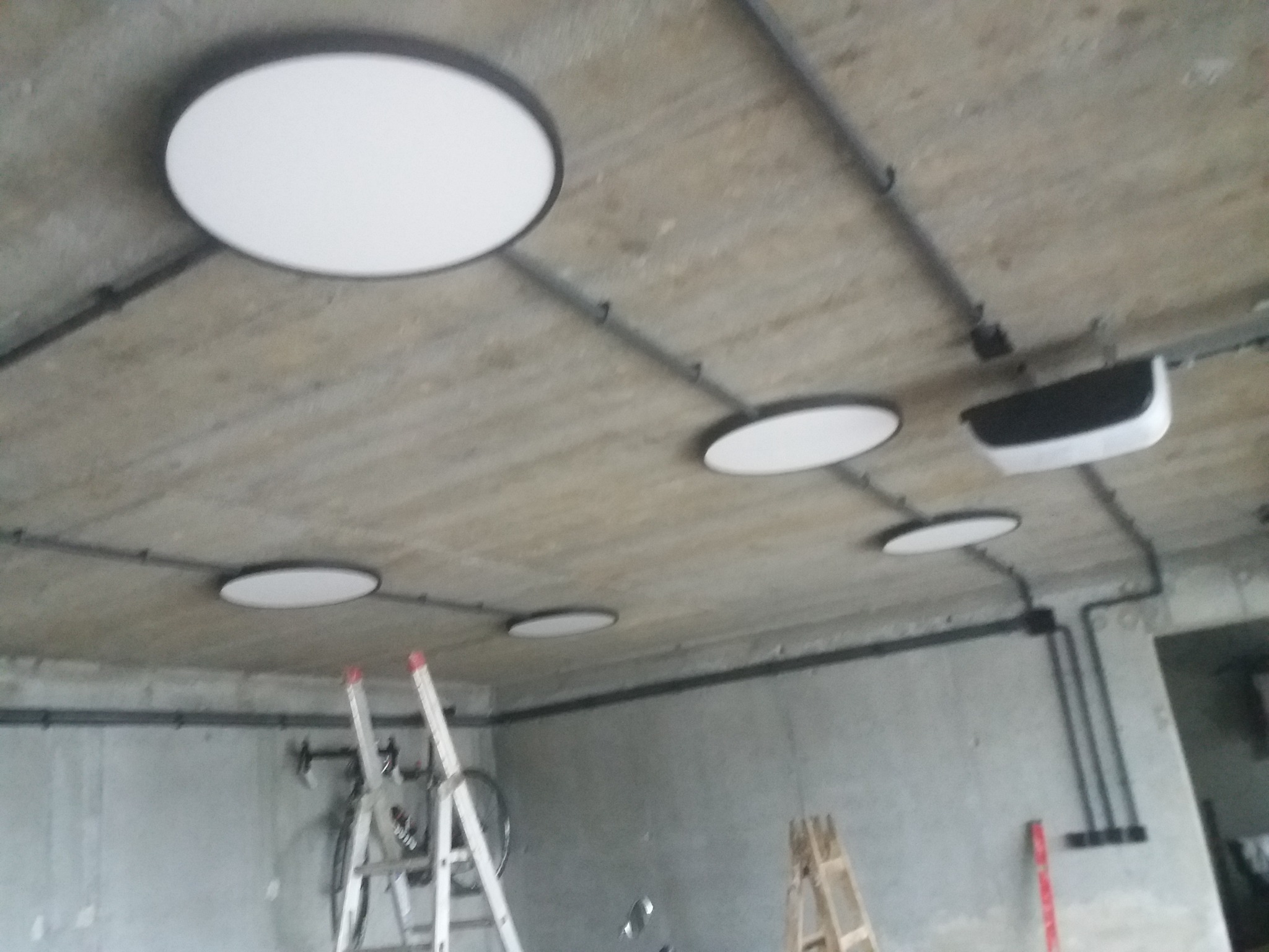 Montaż okrągłych lamp sufitowych w pomieszczeniu z surowym betonowym stropem, widoczne czarne rury instalacyjne i drabiny.
