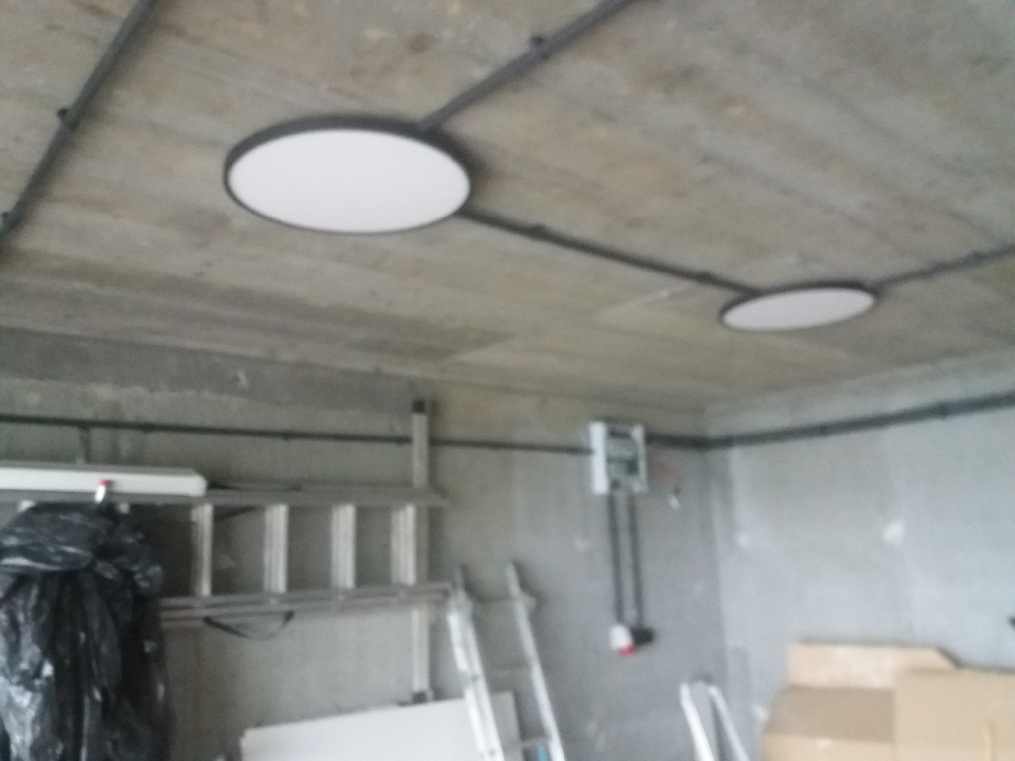 Dwa okrągłe panele LED zamontowane na betonowym suficie garażu z widocznymi przewodami elektrycznymi, drabina i inne akcesoria w tle.