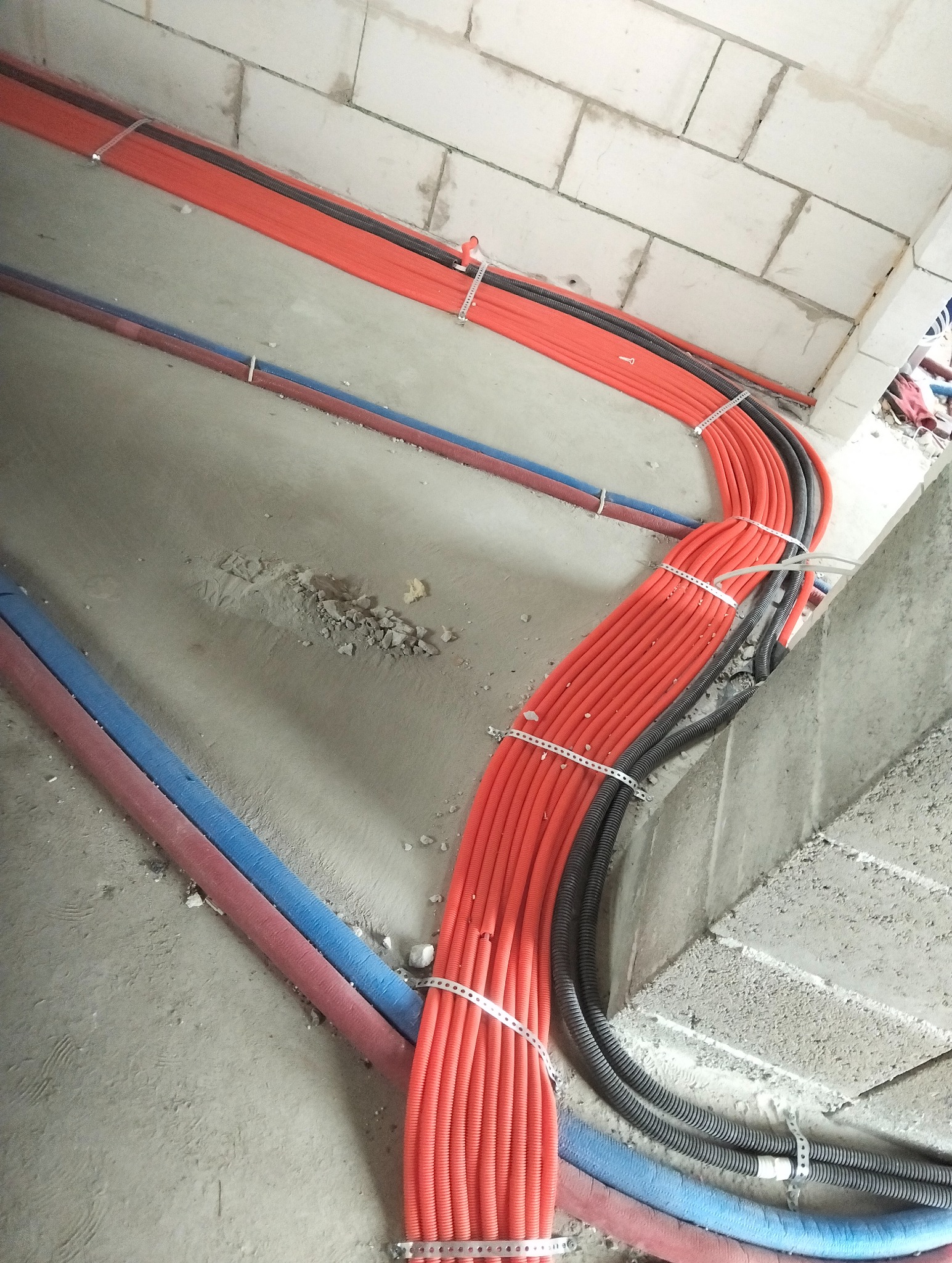 Wiązka pomarańczowych i czarnych rur instalacyjnych, przymocowanych metalowymi obejmami do surowego podłoża, biegnąca wzdłuż ściany z bloczków.