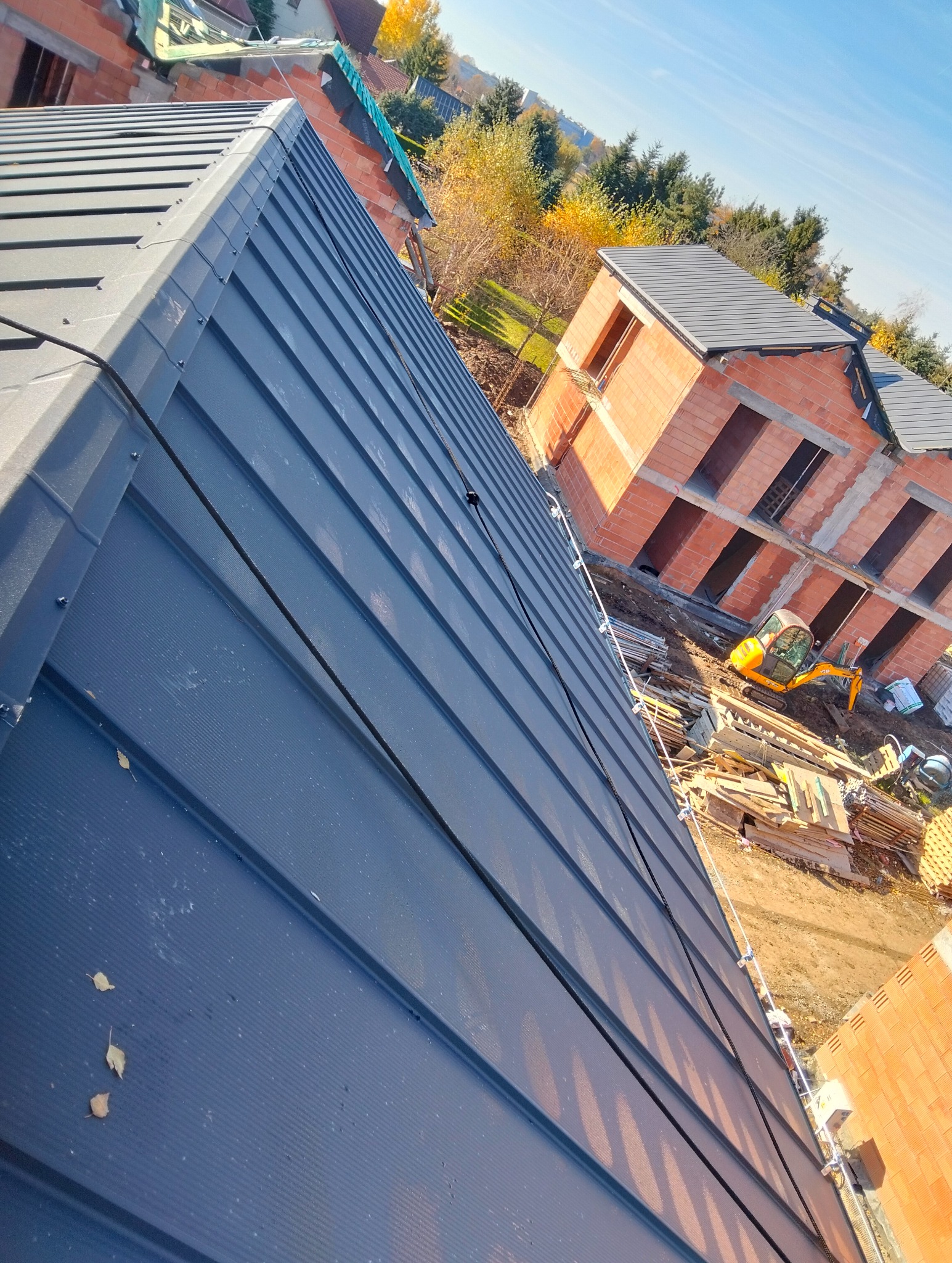 Ciemna blacha na dachu w budowie, widok z góry pod kątem. W tle budynek w stanie surowym i mała koparka. Jesienne liście na dachu.