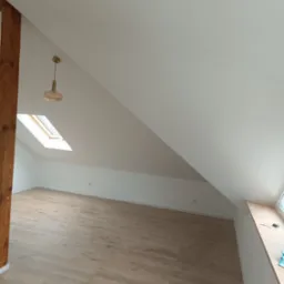 Poddasze po remoncie: jasne panele podłogowe, białe ściany, drewniana belka konstrukcyjna, okno dachowe i wisząca lampa w stylu retro.