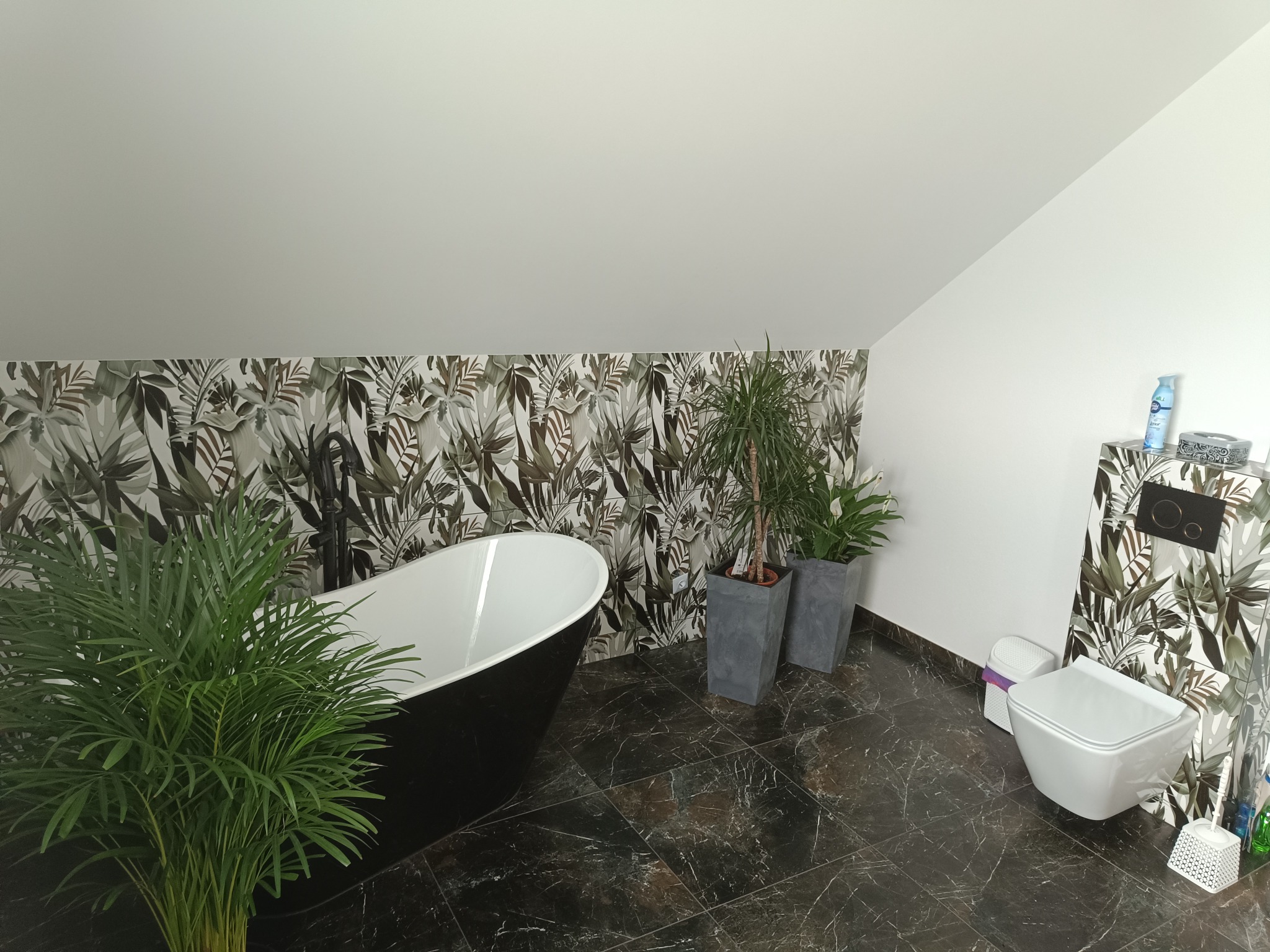 Nowoczesna łazienka na poddaszu z czarną wanną wolnostojącą, marmurową podłogą i roślinami doniczkowymi, ozdobiona płytkami z motywem tropikalnych liści na ścianie.