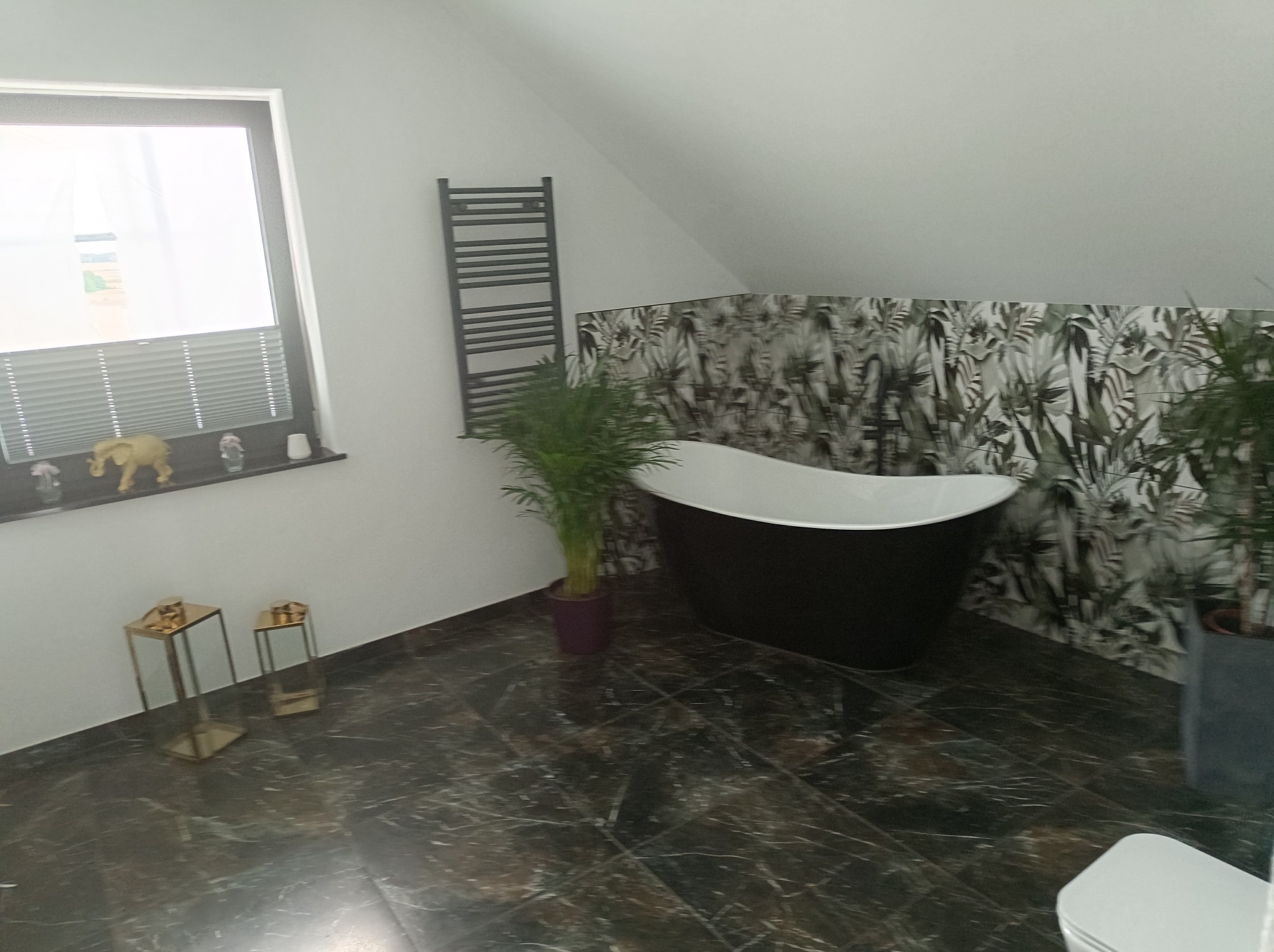 Nowoczesna łazienka na poddaszu z wolnostojącą czarną wanną na tle ściany w tropikalne wzory, ciemna marmurowa podłoga, grzejnik drabinkowy i dekoracyjne złote stoliki pomocnicze.