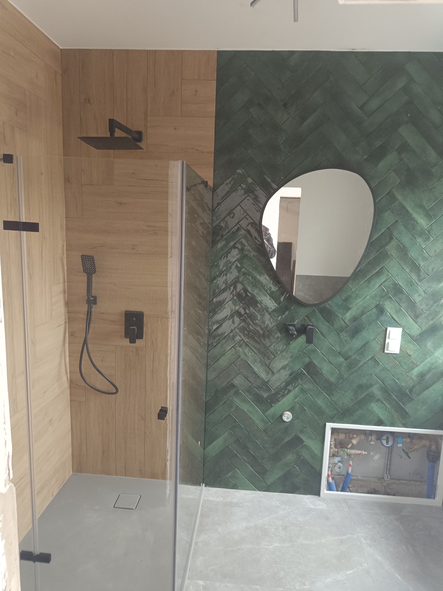 Nowoczesna łazienka z kabiną prysznicową typu walk-in, wyłożona płytkami w dwóch odmiennych stylach: drewnopodobne oraz zielone ułożone we wzór jodełki, czarna armatura, asymetryczne lustro...