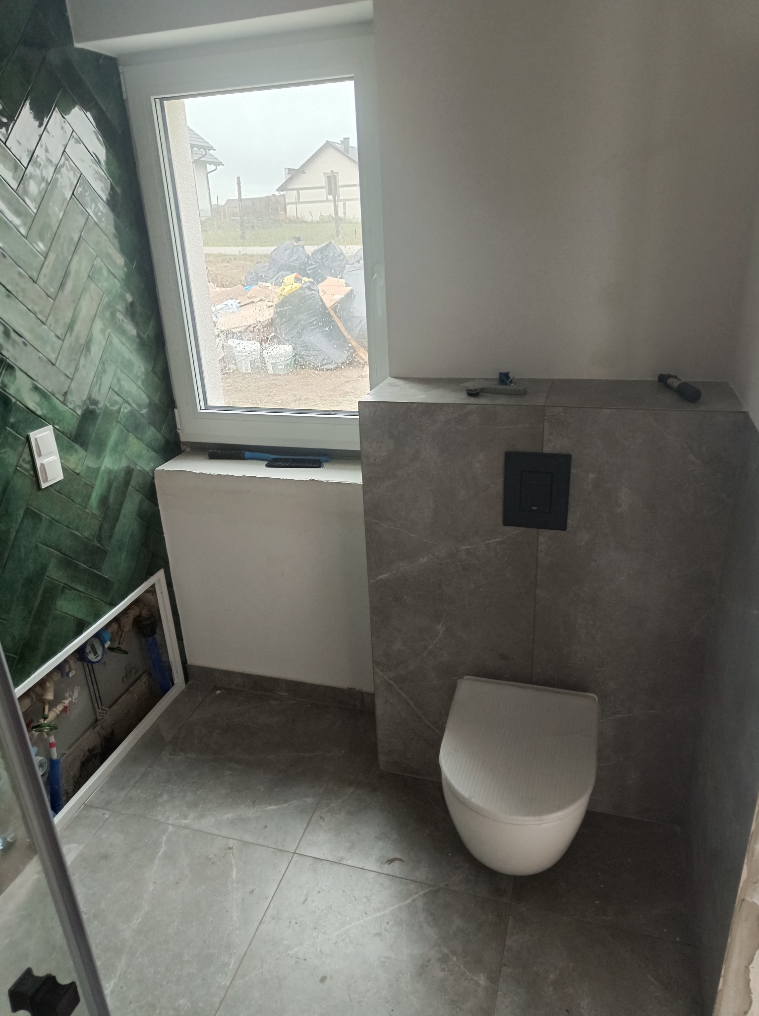 Wnętrze łazienki w trakcie prac: zielona ściana z glazury ułożonej w jodełkę, szara zabudowa stelaża podtynkowego z toaletą, widoczne narzędzia na zabudowie, okno z widokiem na posesję z workami...