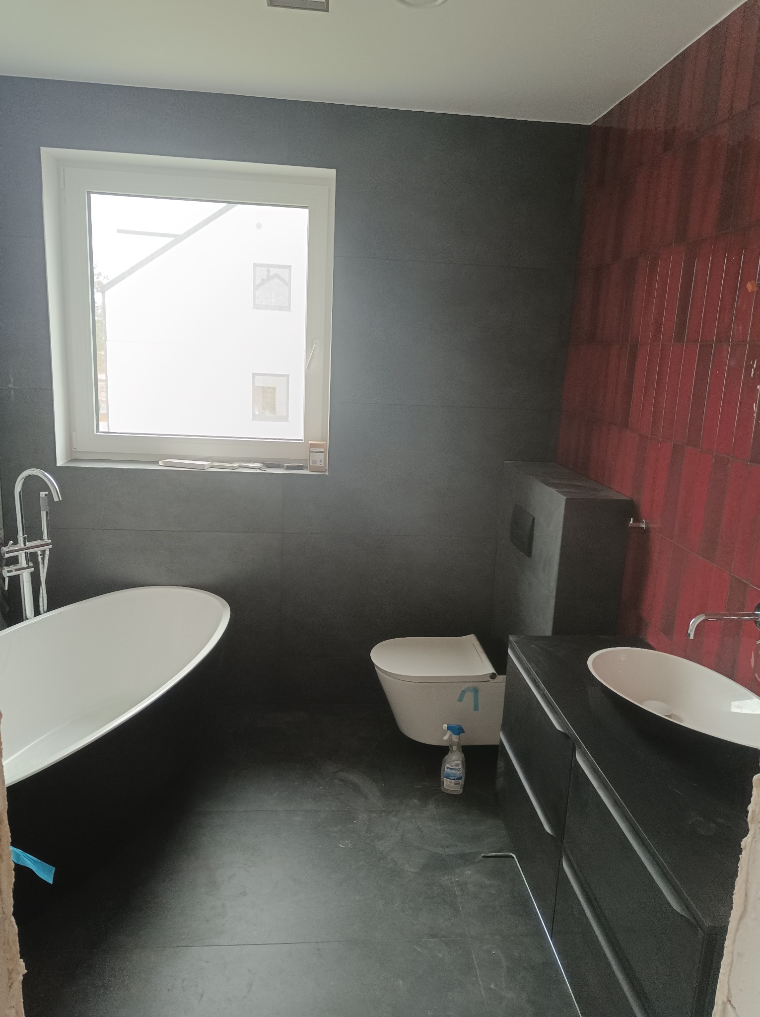 Nowoczesna łazienka z wanną wolnostojącą, umywalką nablatową na szafce z szufladami, toaletą podwieszaną i częściowo wykończoną ścianą w czerwono-szarej mozaice, widoczna butelka środka...