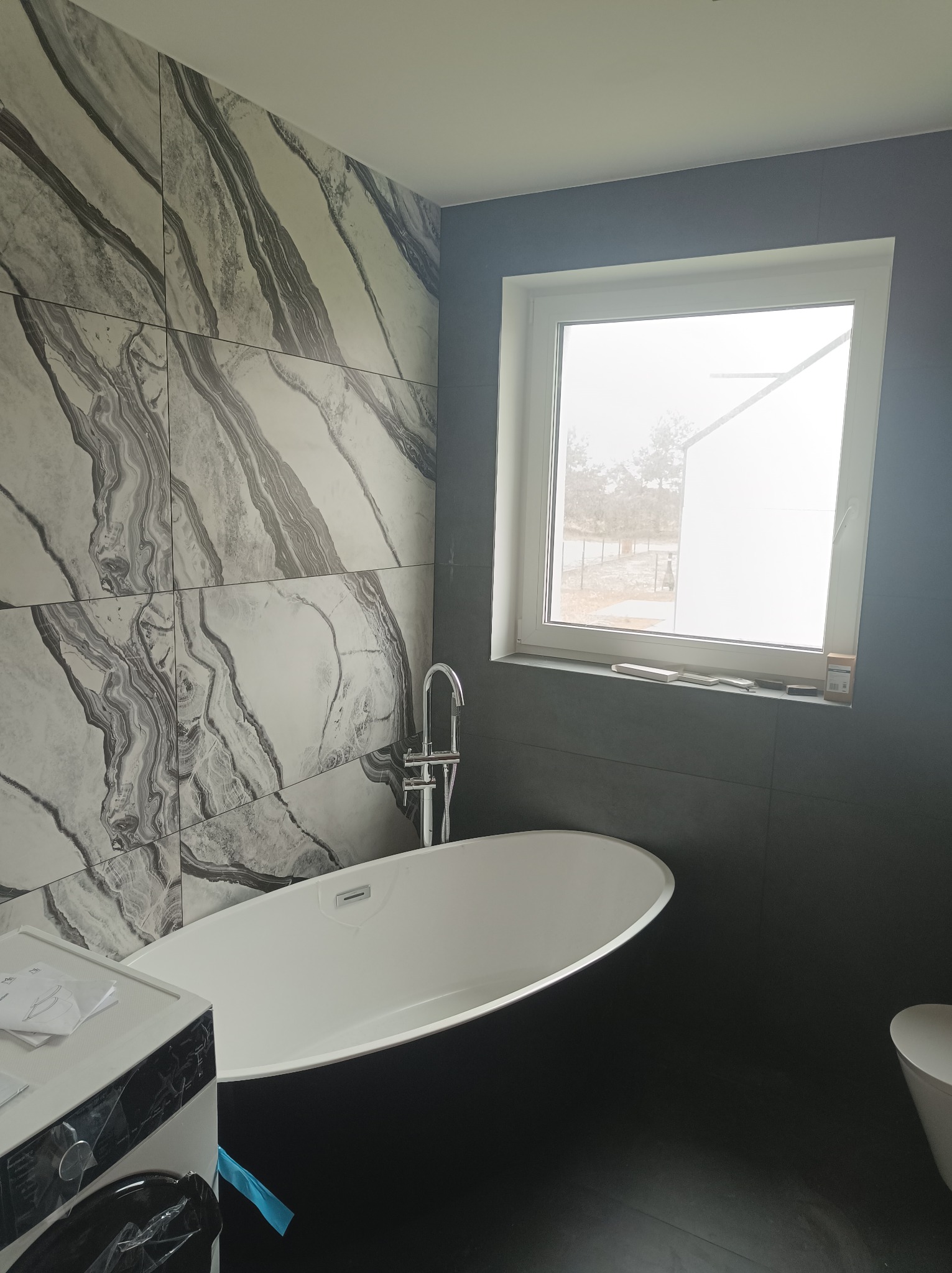 Nowoczesna łazienka z wolnostojącą białą wanną, ściana wyłożona płytkami imitującymi marmur, widok na zewnątrz przez okno, pralka częściowo widoczna po lewej stronie.