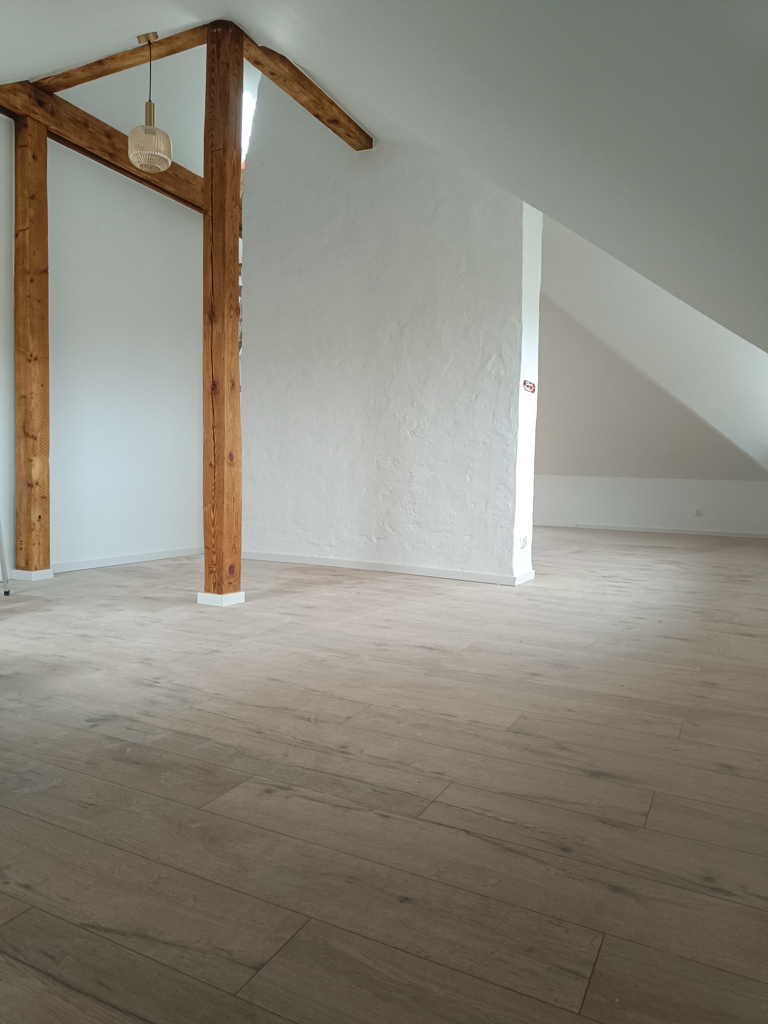 Wnętrze poddasza z drewnianymi belkami konstrukcyjnymi, pomalowanymi na biało ścianami o nierównej fakturze i podłogą wyłożoną panelami imitującymi drewno, oświetlone wiszącą lampą z kloszem...