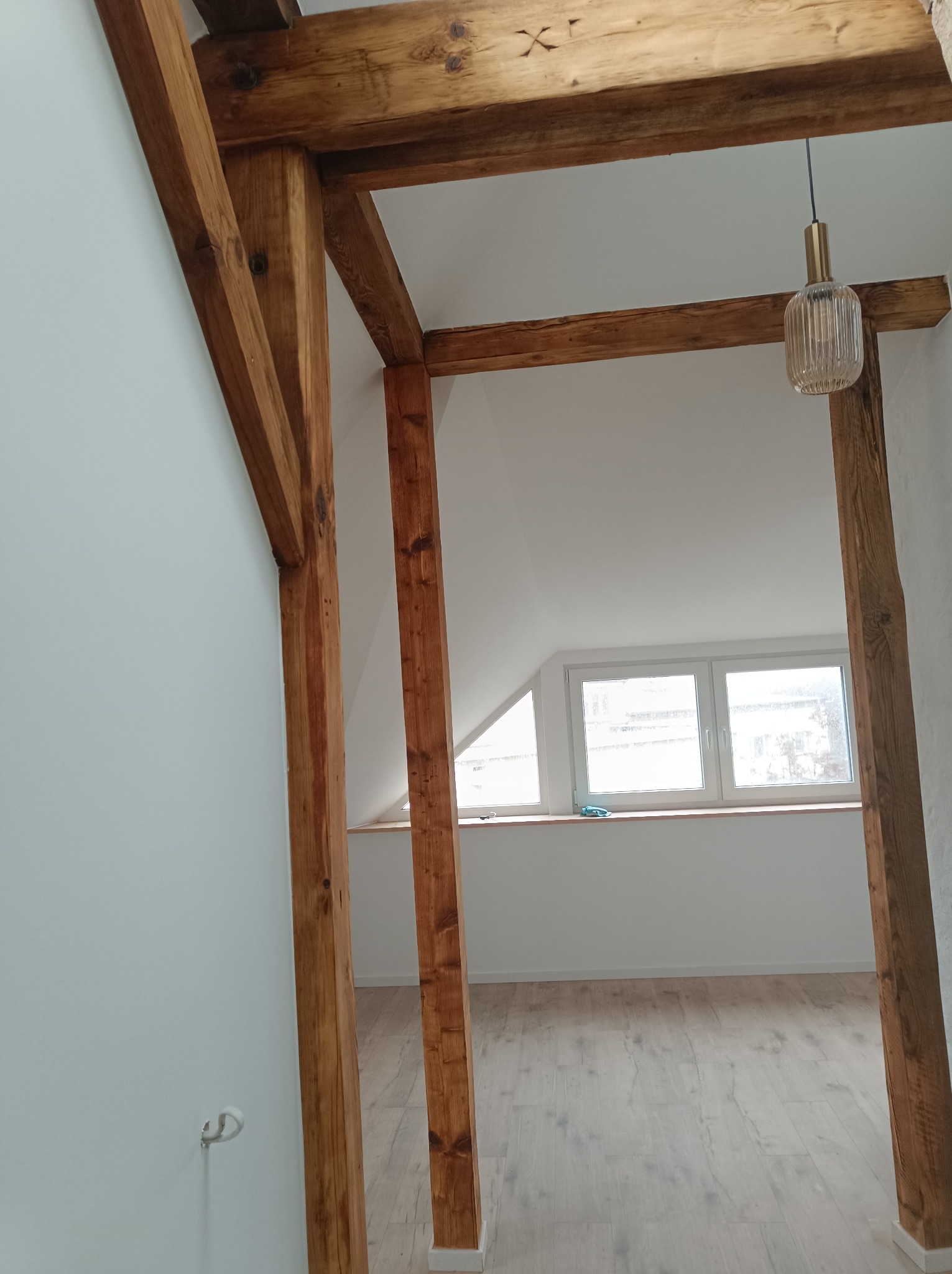 Wnętrze poddasza z drewnianą konstrukcją nośną, świeżo pomalowanymi na biało ścianami i jasną podłogą z paneli, oświetlone wiszącą lampą z żarówką dekoracyjną.