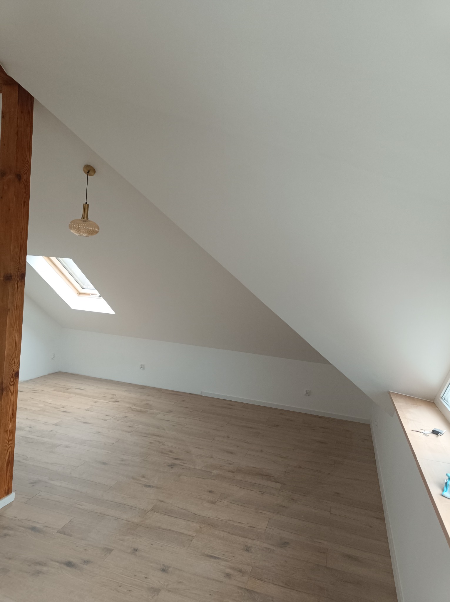 Poddasze po remoncie: jasne panele podłogowe, białe ściany, drewniana belka konstrukcyjna, okno dachowe i wisząca lampa w stylu retro.