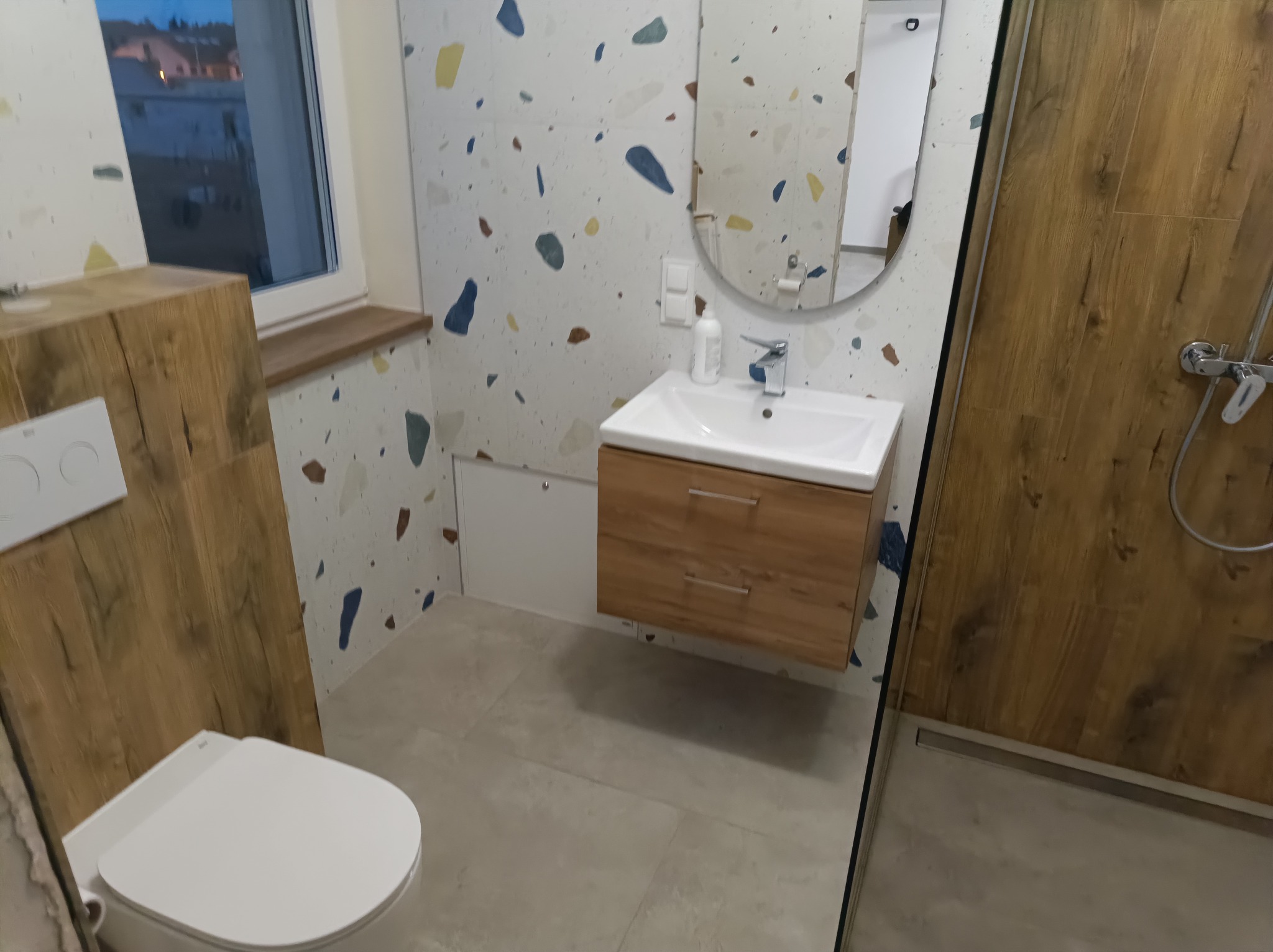 Nowoczesna łazienka z umywalką na szafce z drewnianym wykończeniem, toaleta z deską wolnoopadającą, ściany wyłożone płytkami w stylu lastryko i kabina prysznicowa ze szklanymi drzwiami.