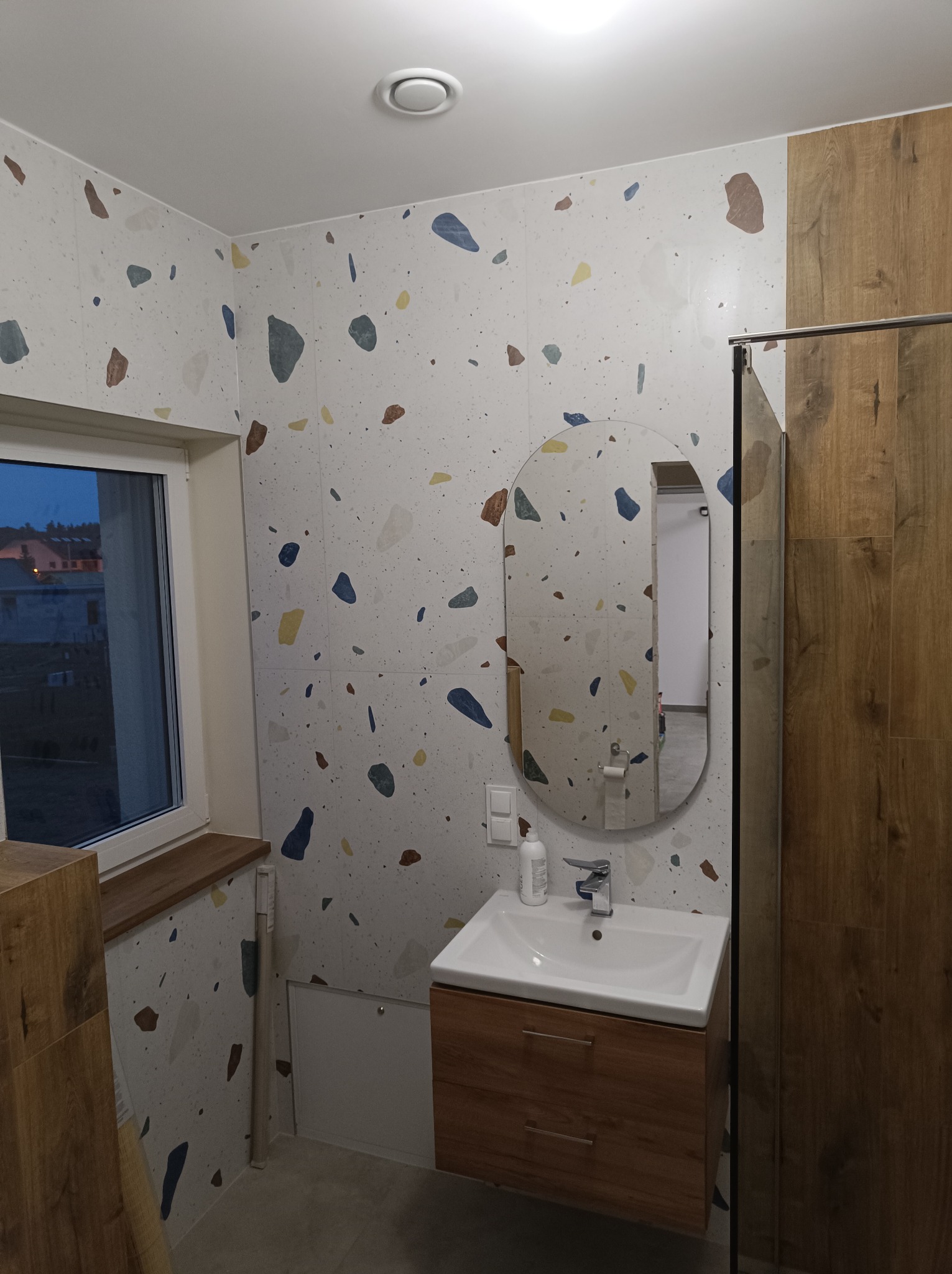 Wykończona łazienka z umywalką na szafce z drewnopodobnym frontem, owalnym lustrem i ścianami w płytkach imitujących lastryko, widok na okno i kabinę prysznicową.