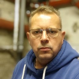 Tomasz Juszczyński TOMI-INSTAL INSTALACJE - Portret mężczyzny w okularach i niebieskiej bluzie z kapturem na tle rur instalacyjnych w pomieszczeniu.