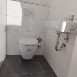 Tomasz Juszczyński TOMI-INSTAL INSTALACJE - Nowoczesna toaleta podwieszana i umywalka w małej łazience z szarymi płytkami podłogowymi i białymi płytkami ściennymi, widoczny przycisk spłukiwania oraz syfon umywalki.