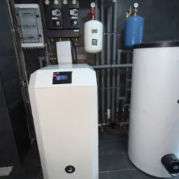 Nowoczesna kotłownia z białym piecem, zbiornikiem buforowym i szarą instalacją rurową na tle ciemnej ściany i szarej podłogi z płytek.