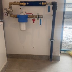 Tomasz Juszczyński TOMI-INSTAL INSTALACJE - Zamontowany filtr wody z niebieską obudową i rurami miedzianymi na białej ścianie, widoczne zawory i manometr. Szare płytki podłogowe.