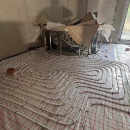 Tomasz Juszczyński TOMI-INSTAL INSTALACJE - Białe rury ogrzewania podłogowego ułożone na folii z czerwoną siatką w surowym wnętrzu. Widoczny fragment osoby w kamizelce odblaskowej i okno w tle.