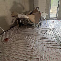 Tomasz Juszczyński TOMI-INSTAL INSTALACJE - Białe rury ogrzewania podłogowego ułożone na folii z czerwoną kratką w surowym wnętrzu. Widoczne materiały budowlane i okno z widokiem na zewnątrz.