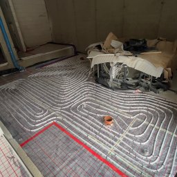 Tomasz Juszczyński TOMI-INSTAL INSTALACJE - Białe rury ogrzewania podłogowego ułożone na czerwonej siatce w surowym wnętrzu. Widoczne elementy instalacji i materiały budowlane. Etap prac przed wylaniem posadzki.