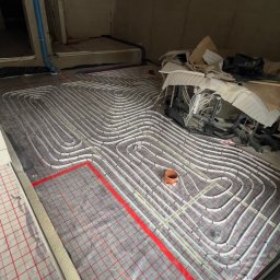 Tomasz Juszczyński TOMI-INSTAL INSTALACJE - Ułożone rury ogrzewania podłogowego na folii z czerwoną siatką, widoczne elementy instalacji i fragmenty ścian w surowym stanie. Pomieszczenie w trakcie prac instalacyjnych.