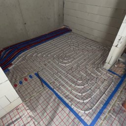 Tomasz Juszczyński TOMI-INSTAL INSTALACJE - Białe rury ogrzewania podłogowego ułożone spiralnie na folii z czerwoną siatką w surowym wnętrzu. Niebieska taśma mocuje rury przy ścianach. Widoczne szare ściany z betonu komórkowego.
