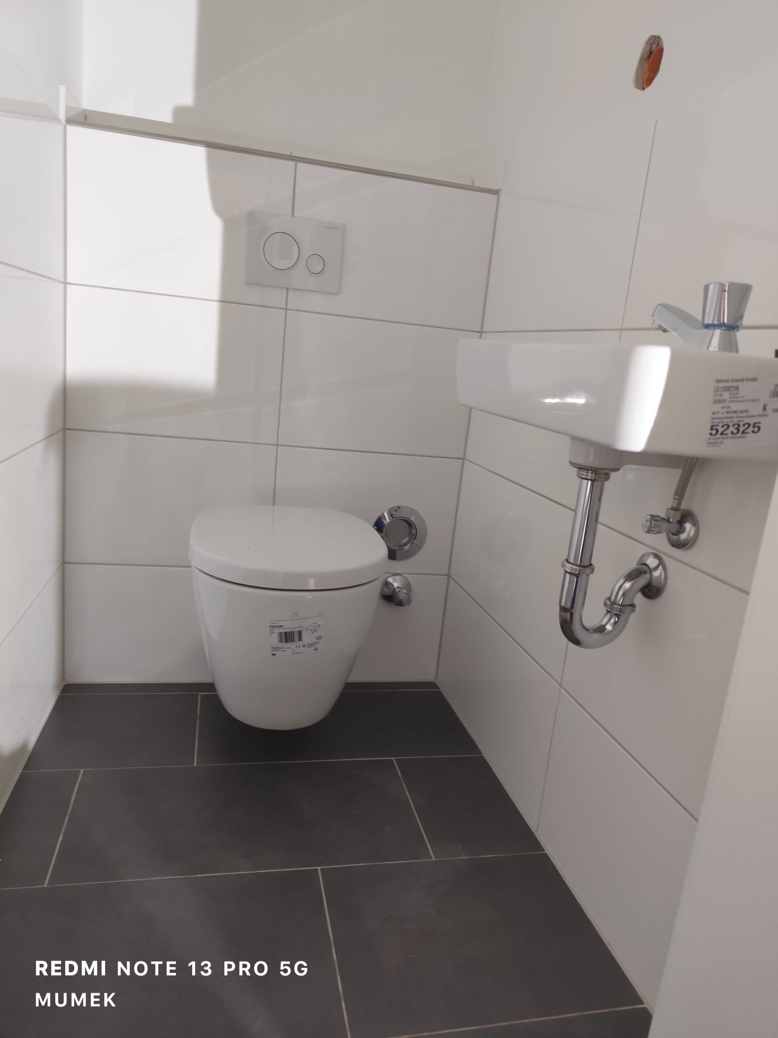 Nowoczesna toaleta podwieszana i umywalka w małej łazience z szarymi płytkami podłogowymi i białymi płytkami ściennymi, widoczny przycisk spłukiwania oraz syfon umywalki.