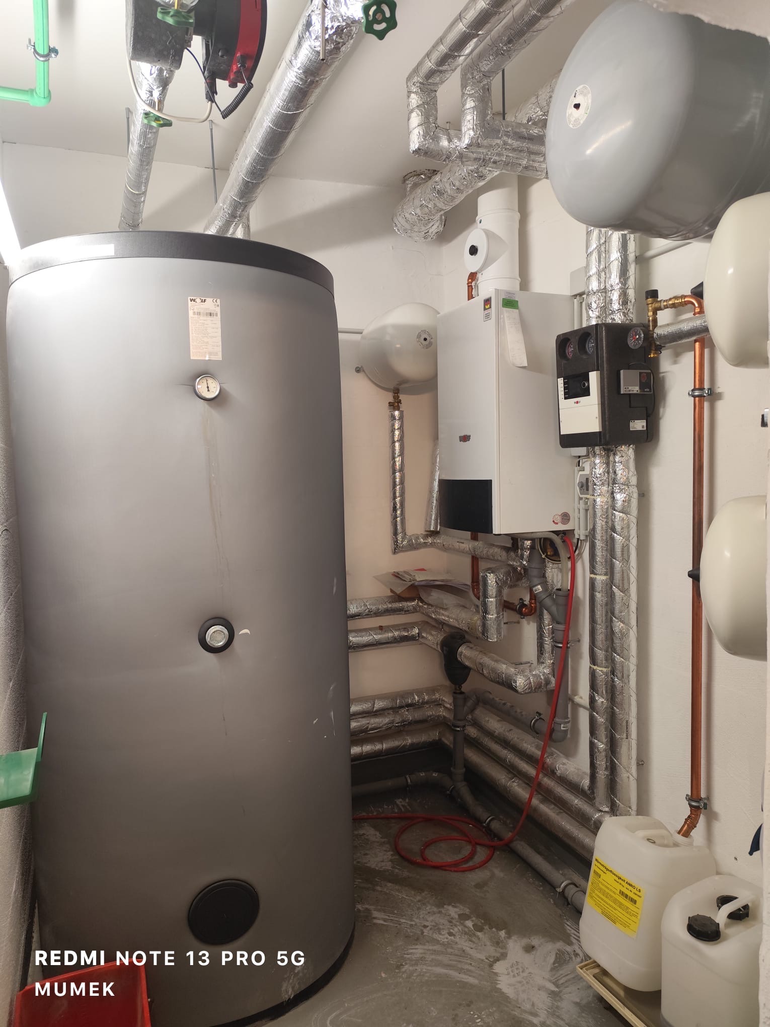 Szary zasobnik ciepłej wody użytkowej z manometrem, obok piec gazowy z czarnym sterownikiem, rury w izolacji aluminiowej, kanistry z płynem grzewczym na posadzce w kotłowni.