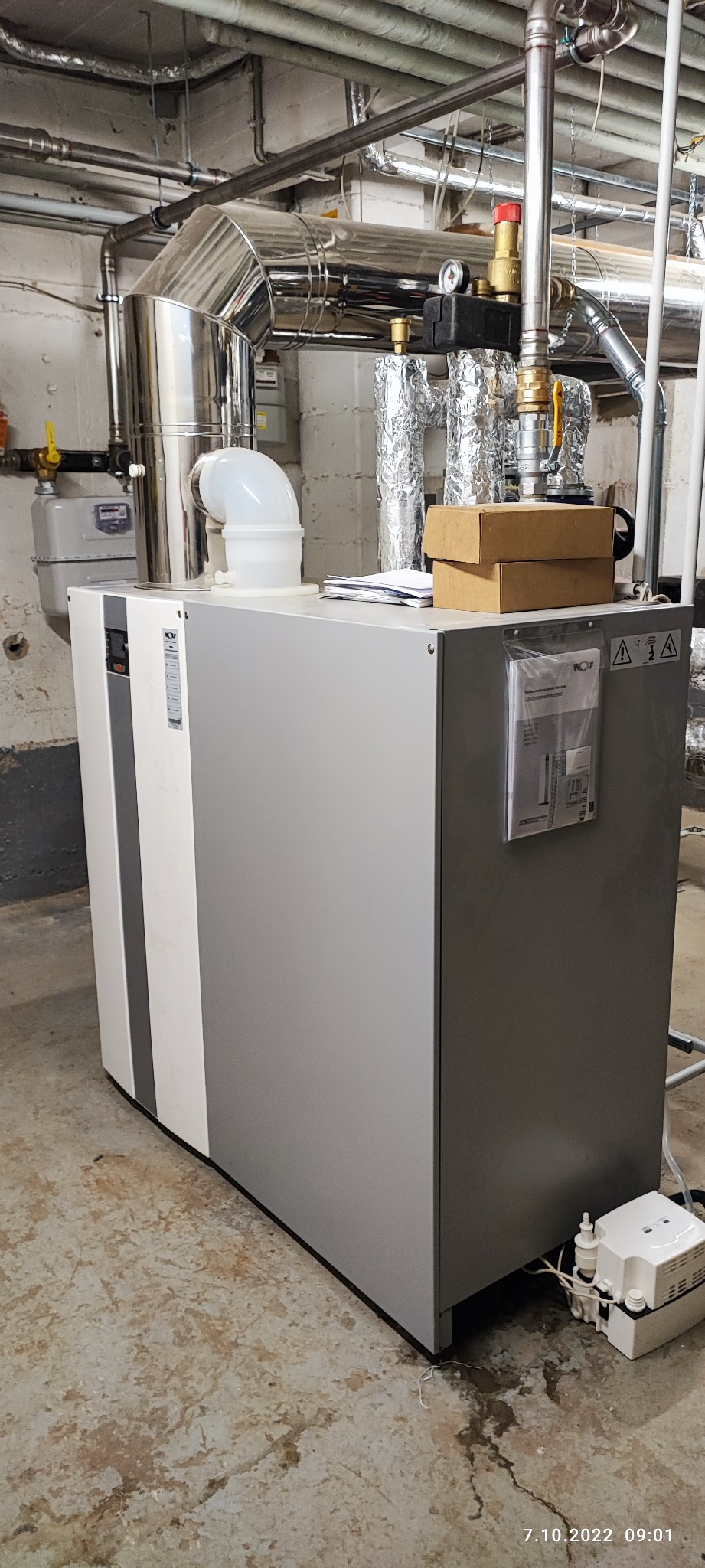 Szary piec gazowy Wolf z białym panelem, widoczne rury instalacji, kondensat na podłodze i instrukcja obsługi w plastikowej koszulce na obudowie.