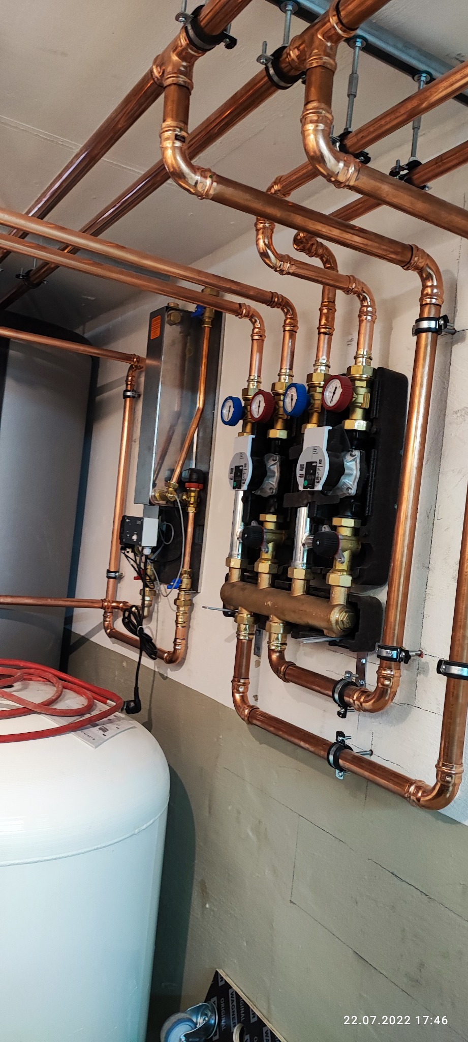 Miedziana instalacja centralnego ogrzewania z rozdzielaczem, pompami obiegowymi i manometrami, zamontowana na ścianie, widoczny zbiornik wyrównawczy i elementy elektryczne
