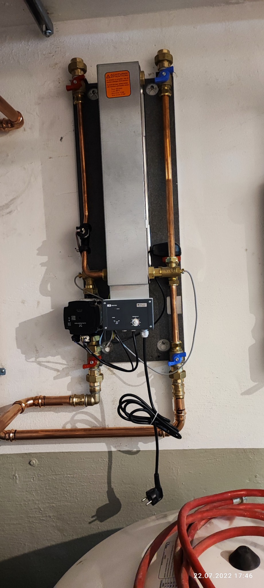 Instalacja centralnego ogrzewania z miedzianymi rurami, pompą obiegową Grundfos i regulatorem temperatury, zamontowana na ścianie z widoczną instalacją elektryczną i fragmentem zbiornika...