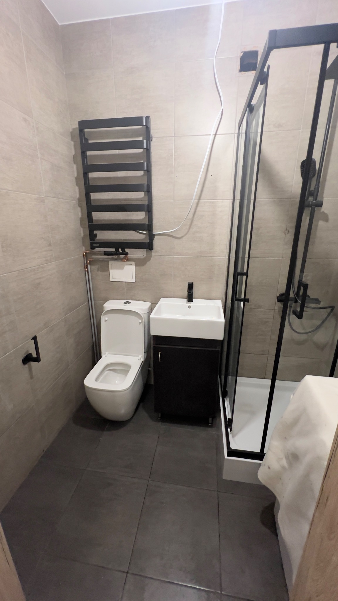 Nowoczesna łazienka z kabiną prysznicową w czarnej ramie, wiszącą szafką z umywalką, toaletą i grzejnikiem drabinkowym w kolorze czarnym. Widoczne rury doprowadzające wodę do grzejnika i elementy...