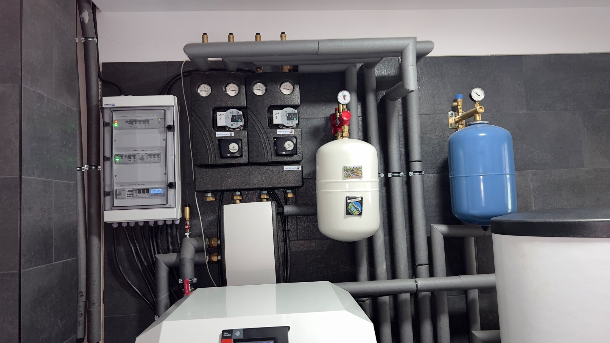 Węzeł cieplny z osprzętem: rozdzielacze, pompy, zawory, zbiorniki ciśnieniowe, automatyka, orurowanie w szarej izolacji, zamontowany na tle ciemnoszarych płytek ściennych.