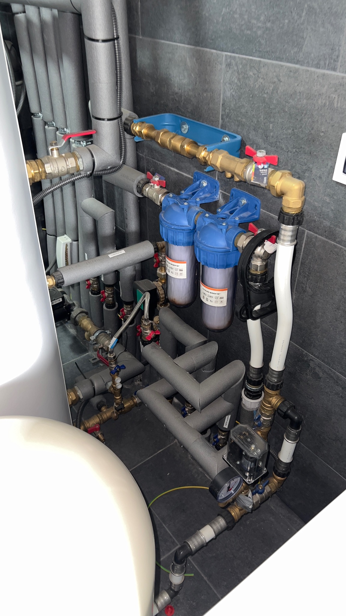Złożony system rur, zaworów i filtrów wody w pomieszczeniu technicznym, większość rur zaizolowana szarą pianką, widoczne dwa niebieskie filtry wody z przezroczystymi obudowami, manometr i elementy...