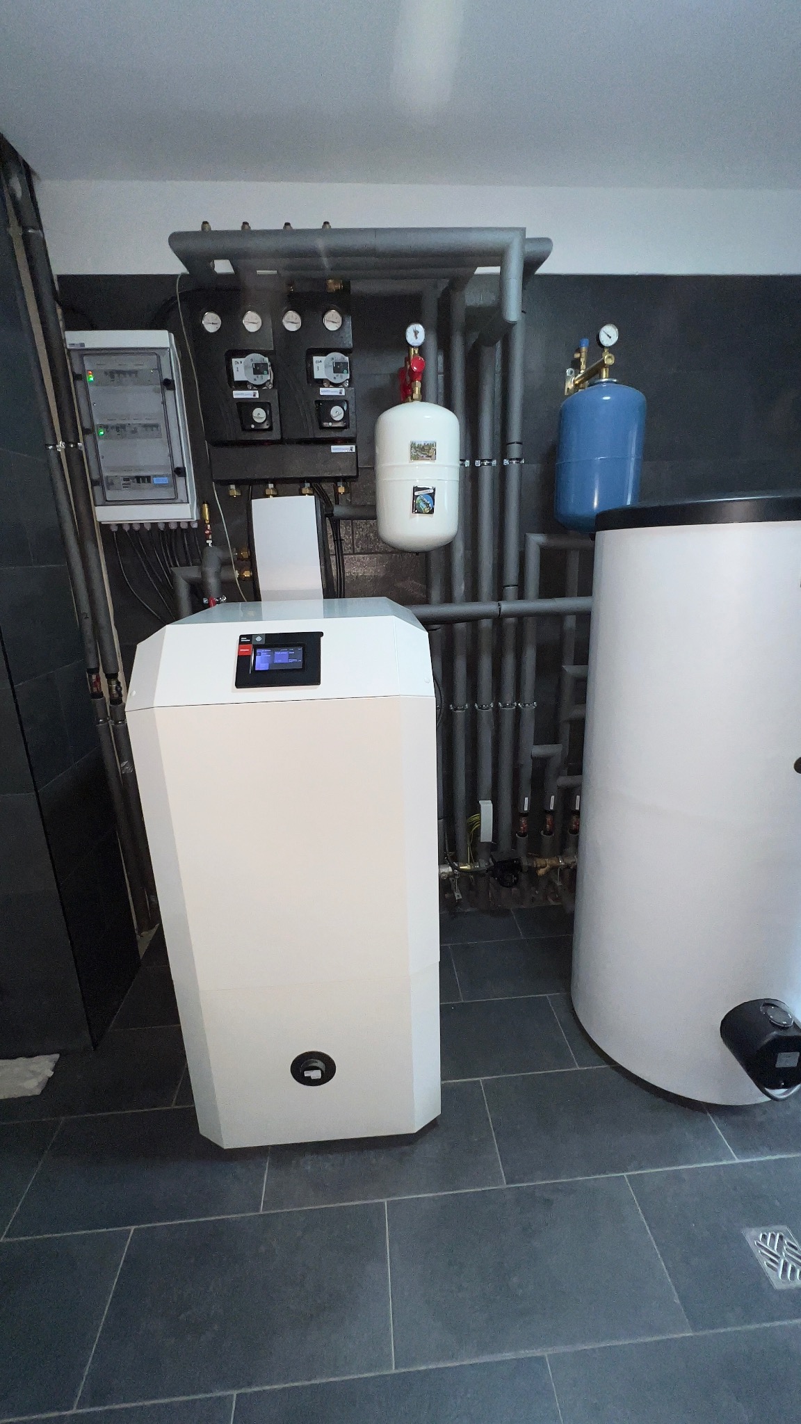 Nowoczesna kotłownia z białym piecem, zbiornikiem buforowym i szarą instalacją rurową na tle ciemnej ściany i szarej podłogi z płytek.