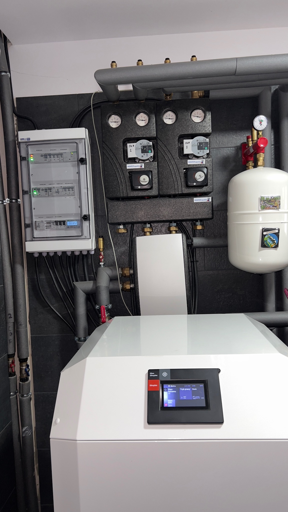 Biały piec grzewczy z czarnym panelem sterowania dotykowego, obok rozdzielnica elektryczna i instalacja grzewcza z rurami w izolacji, manometrami i zaworami na tle ciemnych płytek.